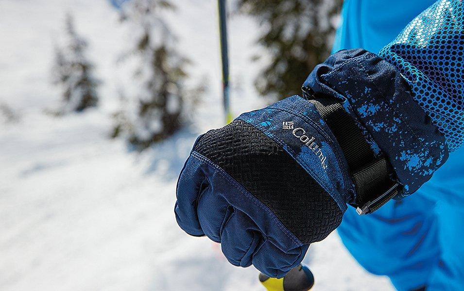 A close-up shot of a blue Columbia Sportswear glove gripping a ski pole in a snowy mountain setting. 