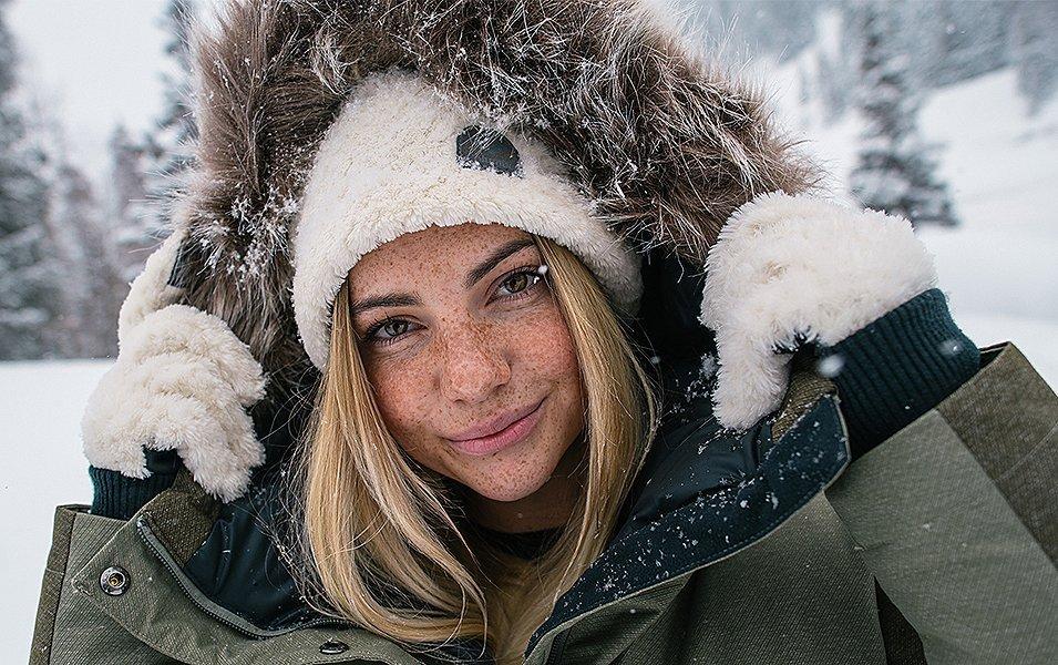 A woman stands in the snow wearing fluffy gloves and a jacket with a faux fur trim smiling at the camera. 