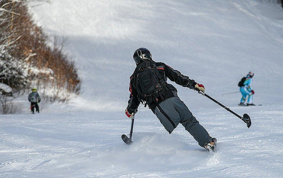 An amputee skier using a single ski and adaptive poles glides down a snow-covered slope, while an instructor watches from a distance.