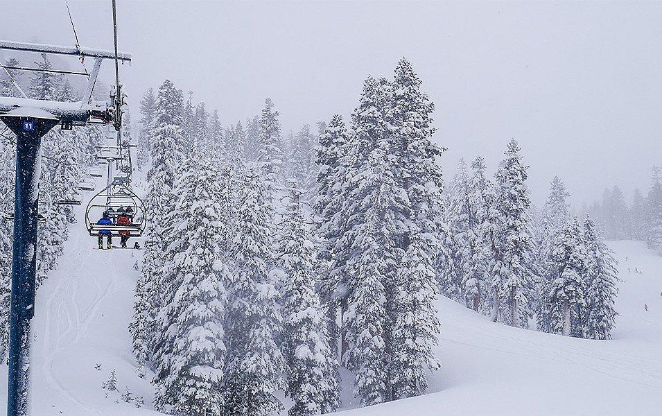 A snowboarder and a skier ride side by side on a chairlift at Mammoth Mountain, passing snow-covered trees in the alpine landscape.