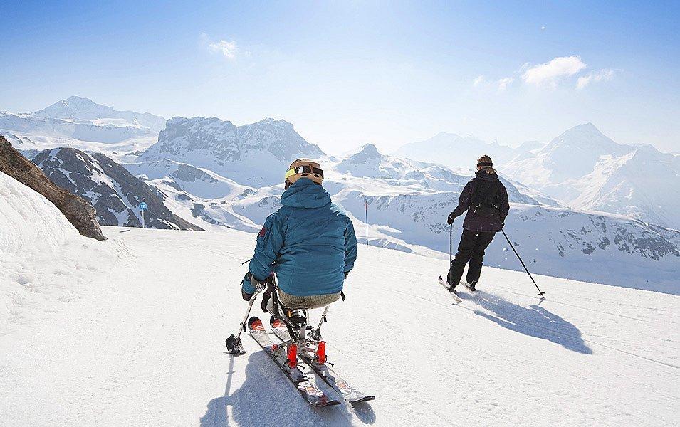 A person using an adaptive sit ski and poles glides down a snow-covered slope alongside a traditional skier. Snow-covered peaks rise in the distant landscape.