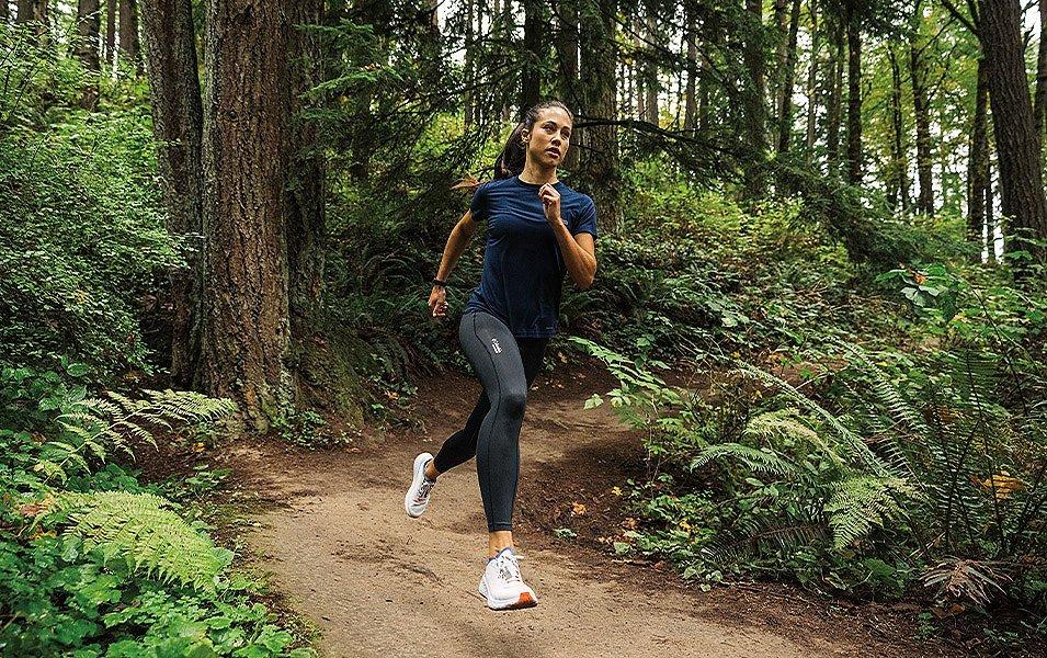 A woman in black tights and a dark blue running T-shirt hits her stride, charging down a narrow dirt trail surrounded by ferns and cedars.