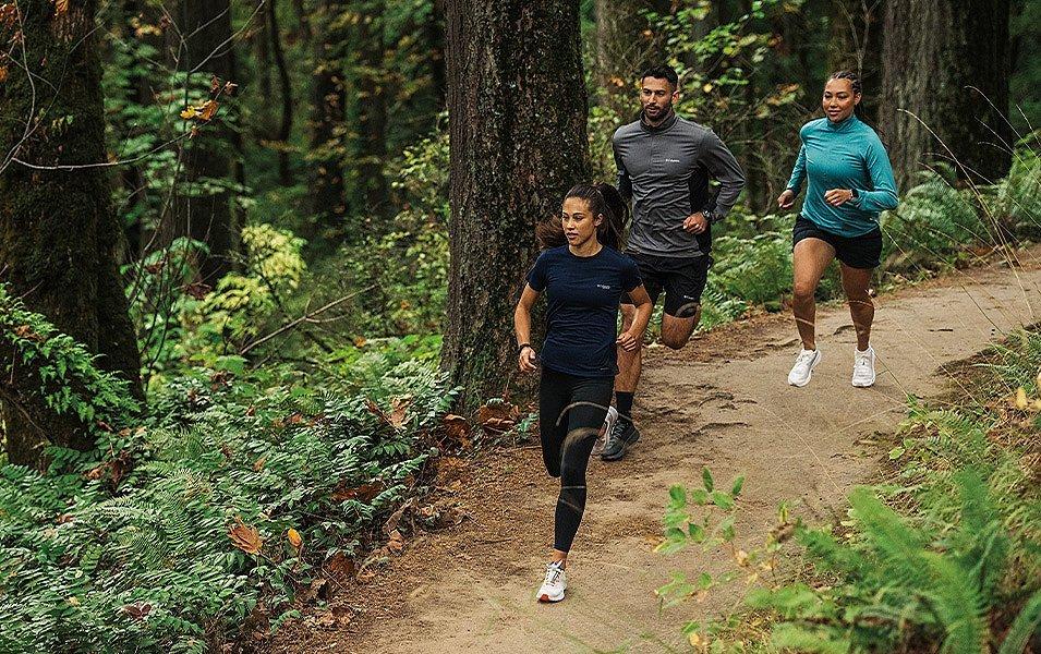 Three runners—two in shorts and one in tights—pick up the pace as they sprint through a forest trail.