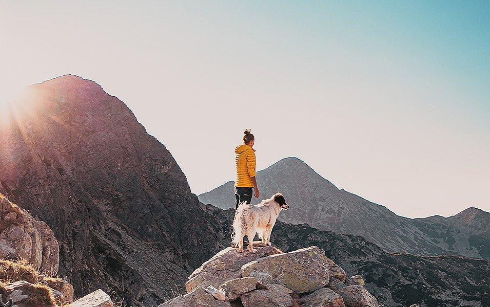 A woman, bundled in a cozy light puffy jacket, stands proudly atop a massive boulder, sharing a quiet moment with her dog as they take in the breathtaking, rugged landscape ahead.