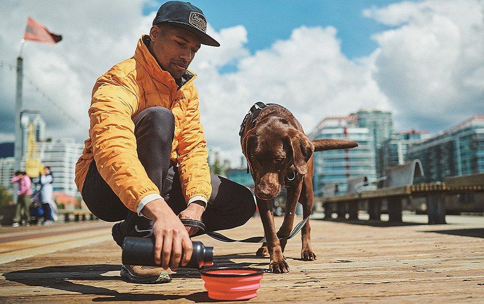 A man fills a portable dog bowl with water from a flask, ensuring his loyal companion stays hydrated on their outdoor journey.