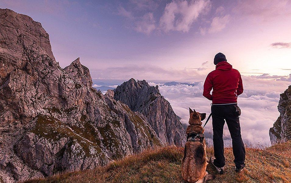 A hiker, with his dog on a leash for safety, pauses to enjoy the view of a vast, mist-covered valley.