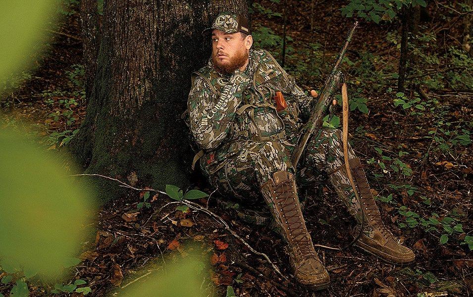 Luke Combs, tests the padded back panel of the Luke Combs Turkey Hunting vest as he leans against a tree, shotgun in hand, scanning for wild turkeys.