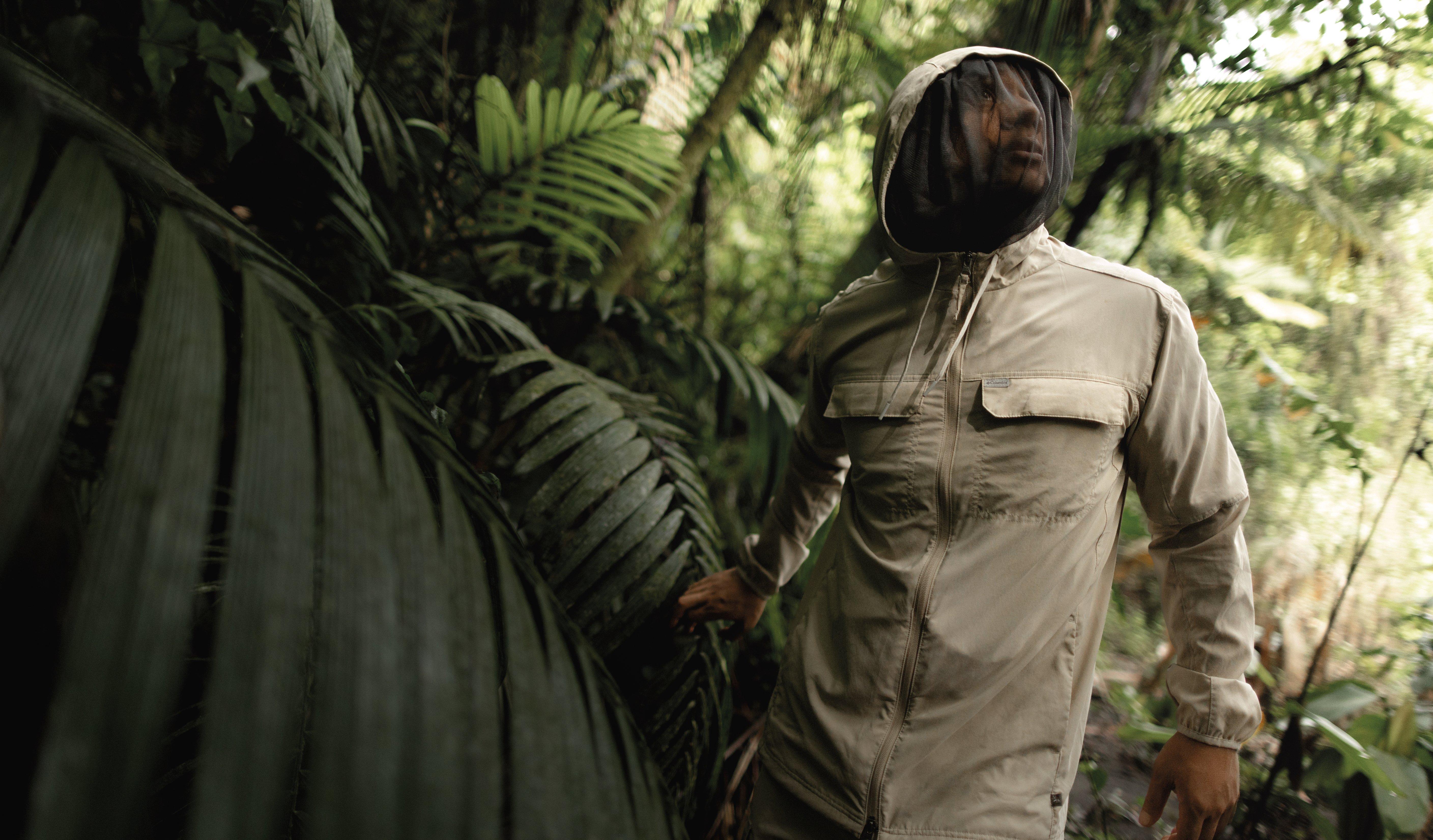 Person wearing beige safari jacket with mesh face protection walking through tropical jungle vegetation.