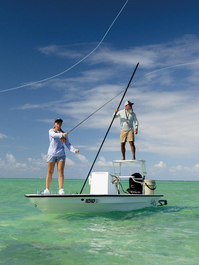 Two people fly fishing from a small boat in clear turquoise waters, one casting while another stands on platform.