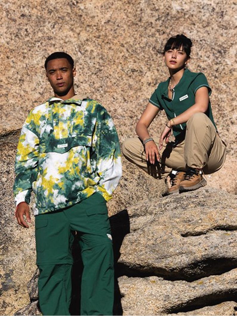 Two models posing on rocky terrain wearing green and yellow tie-dye sweatshirt with green pants and polo.