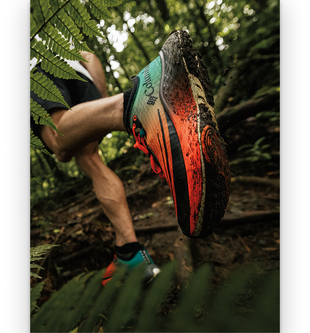 Close-up of muddy orange and black trail running shoe on runner's foot traversing forest terrain.