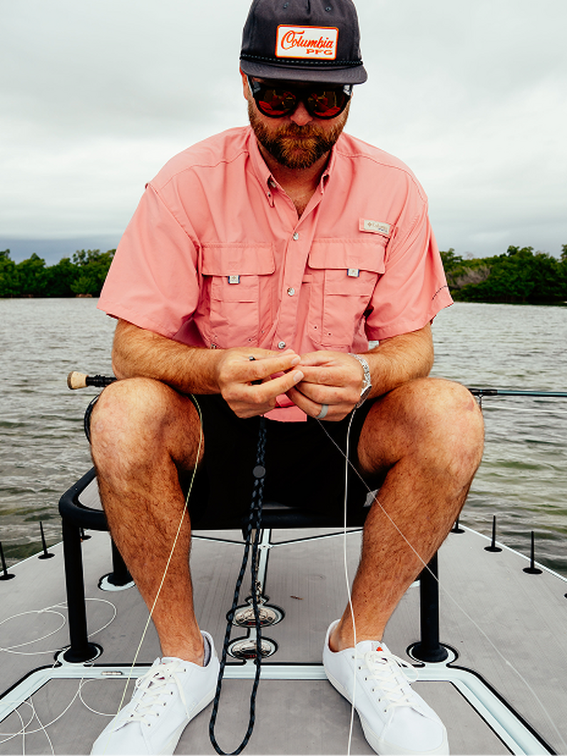 Man sitting on boat wearing coral fishing shirt, black shorts, cap and sunglasses, preparing fishing line.