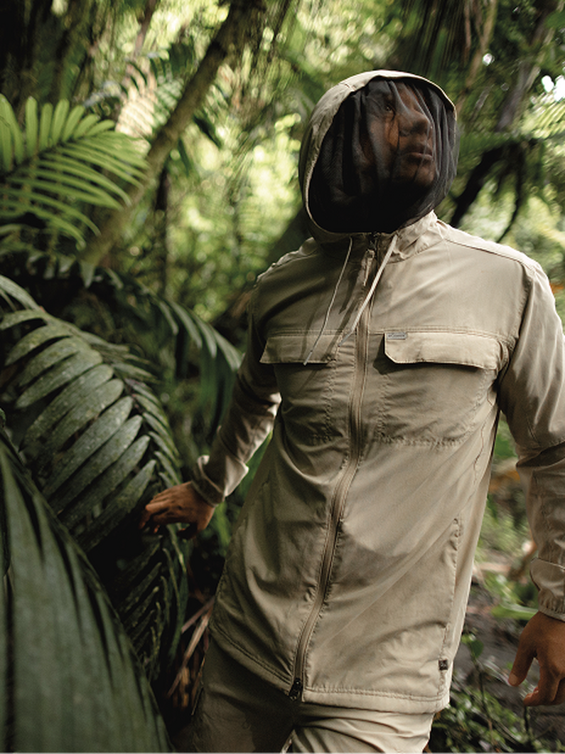 Person wearing tan utility jacket with mesh hood standing in lush tropical forest with ferns.