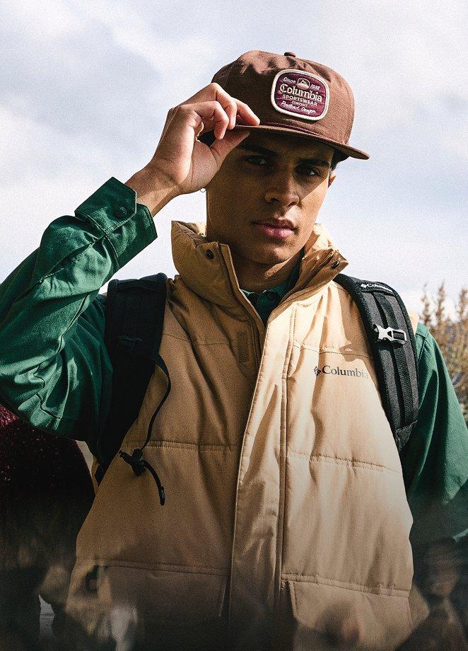 A man hiking on a cloudy day wearing a hat and vest.