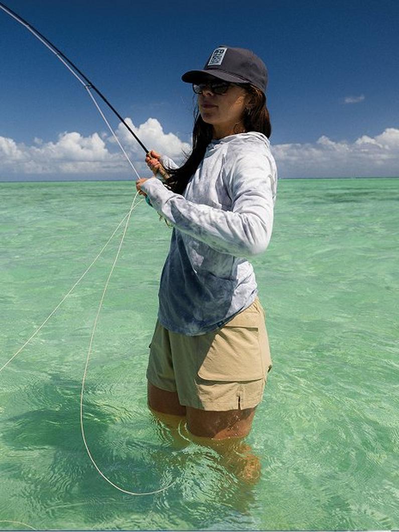 Woman fly fishing in shallow turquoise water wearing long-sleeve shirt, shorts, cap, and sunglasses.