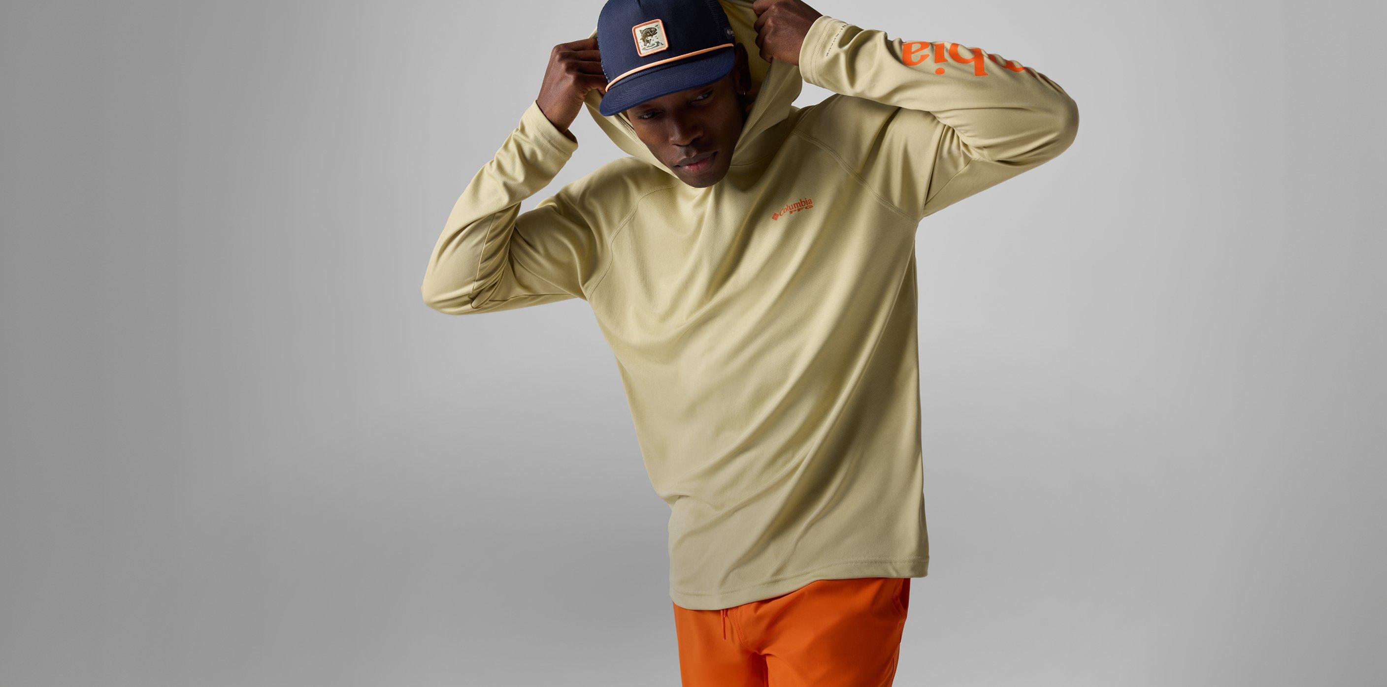 Man adjusting navy blue snapback cap while wearing pale yellow long-sleeve shirt and orange pants.