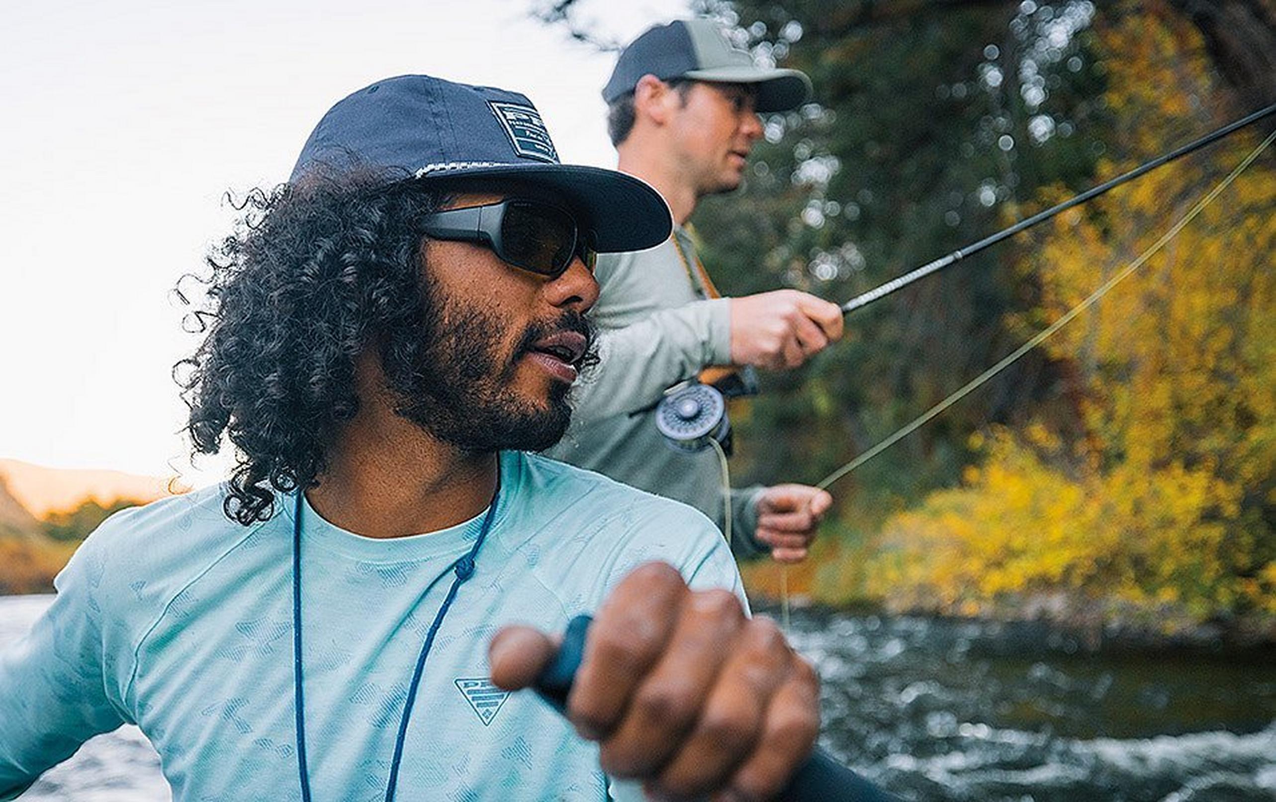 Man in a blue pfg shirt rowing a boat while a person in the background fishes