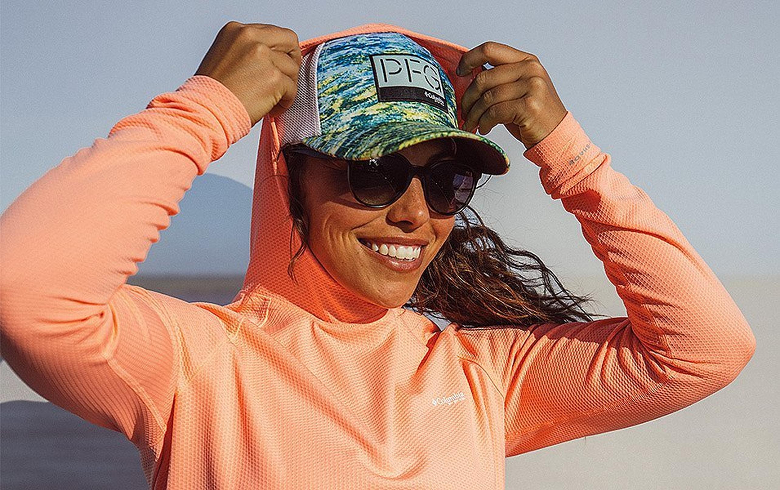 Women wearing a hat and sunglasses pulling an orange upf hood over her head 