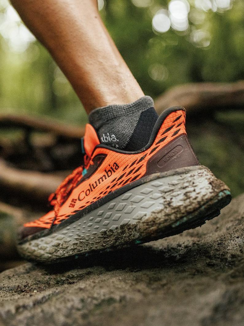 Close-up of model trail running with orange upper Konos shoe. Mud is caked on the bottom of the outsole and Omni-Max cushioning.