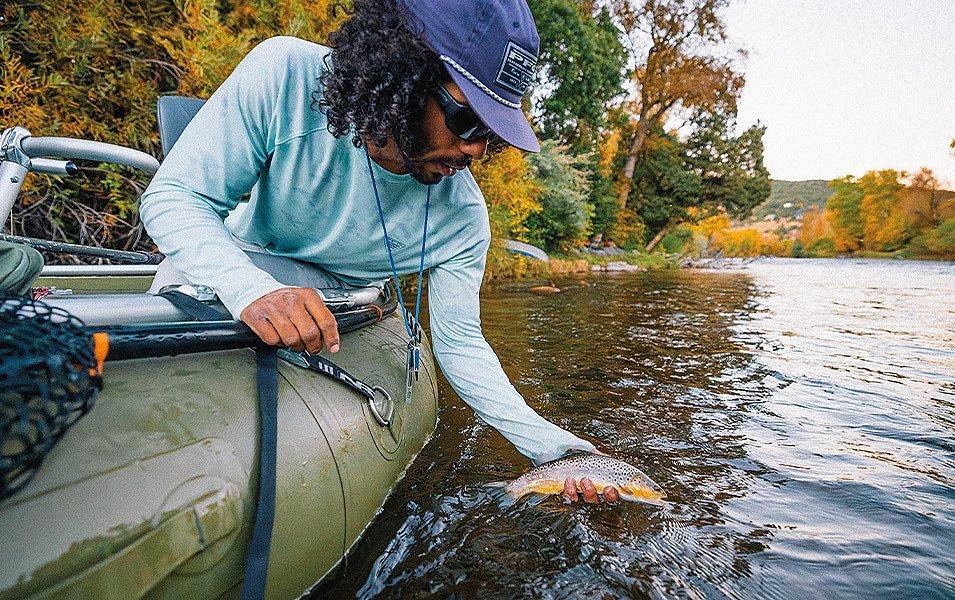 A man wearing Columbia PFG apparel releases a small fish back into the water from the edge of a boat.
