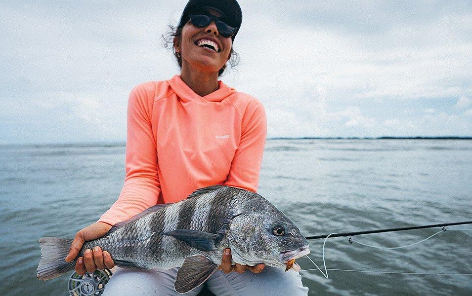 A woman wearing Columbia PFG apparel holding a large striped fish and a fly rod. 