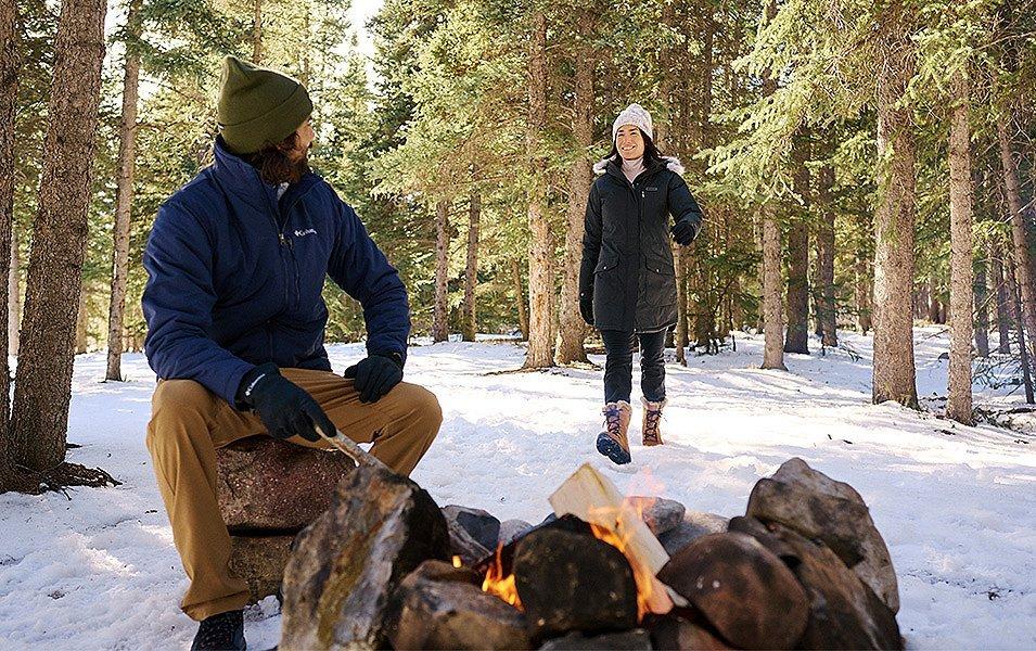 A bearded man stokes an outdoor fire as a woman joins him for an aprés ski celebration. 