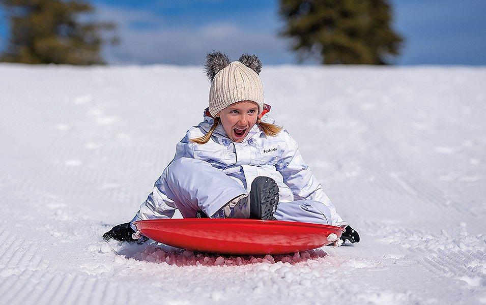 A young girl wearing a knit hat with bunny ears laughs as she rides a red sled down a groomed slope.