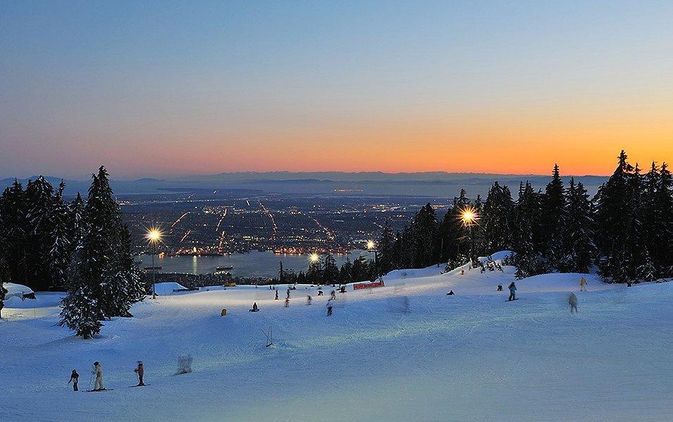 People are skiing with the sun setting in the background. In the distance, you can see city lights.