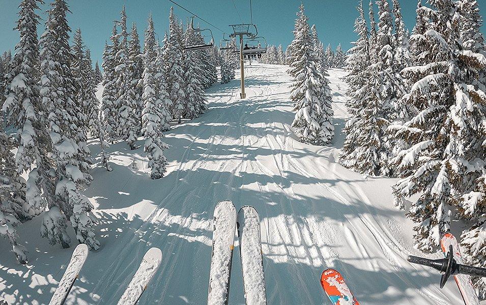 Three skiers are riding up a chairlift. Between their ski tips you can see a snow-laden slope with deep tracks. 