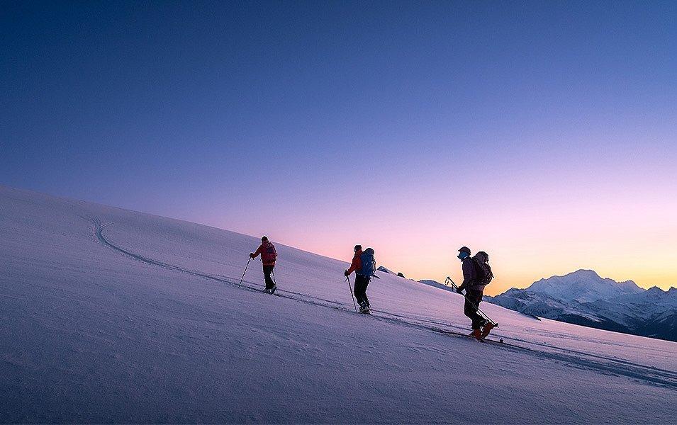 Three skiers with alpine touring gear begin their ascent up a peak as the sun rises.