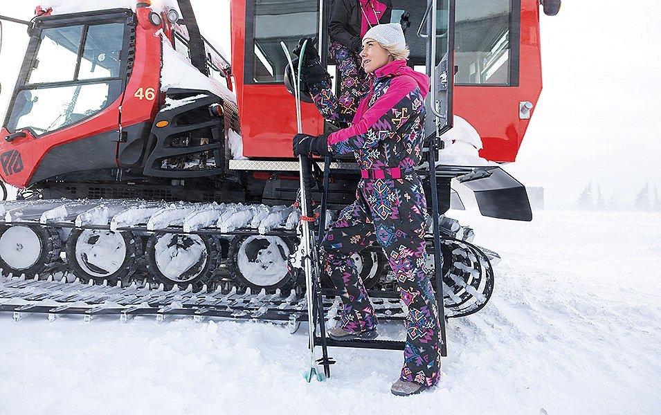 A well-outfitted skier wearing a brightly colored one-piece suit, steps off the snowcat, skis in hand.