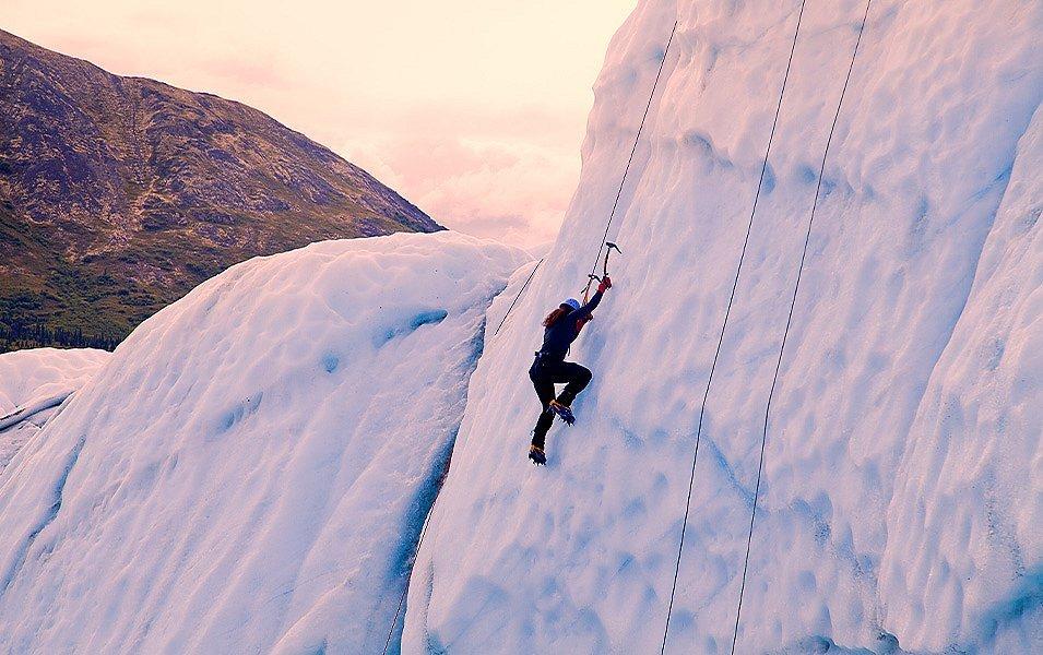 A woman climbs a glacier in the morning light, which casts a pink and lavender hue on the ice. Her perfect technique is supported by crampons, ice axes, and a top rope securing her ascent. 