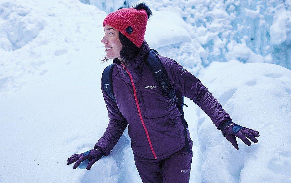A woman in a warm Columbia jacket, pants, and gloves hikes through snow to the base of a frozen waterfall.