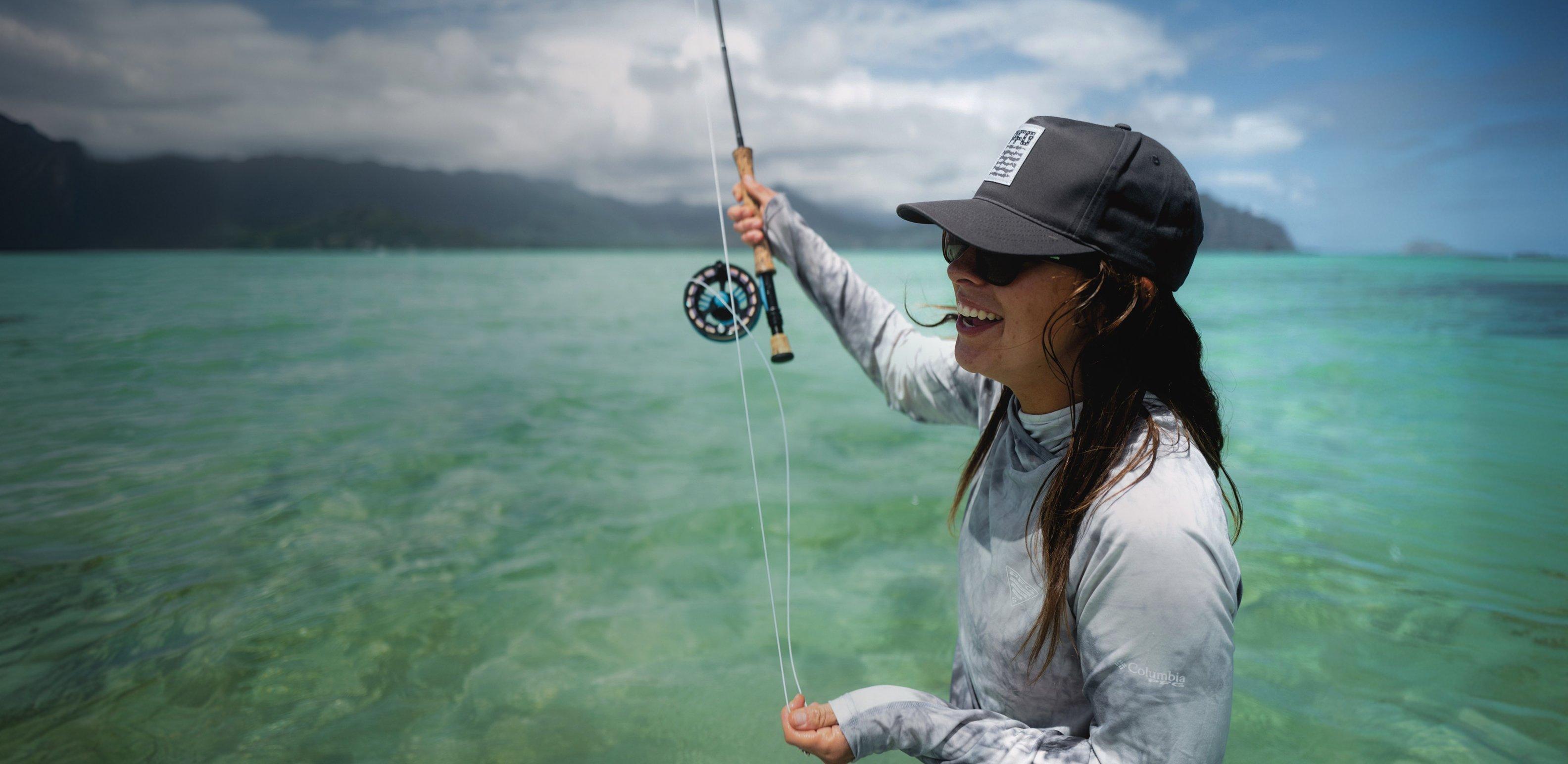 Woman fly fishing in turquoise water wearing dark cap, sunglasses, and long-sleeve fishing shirt.
