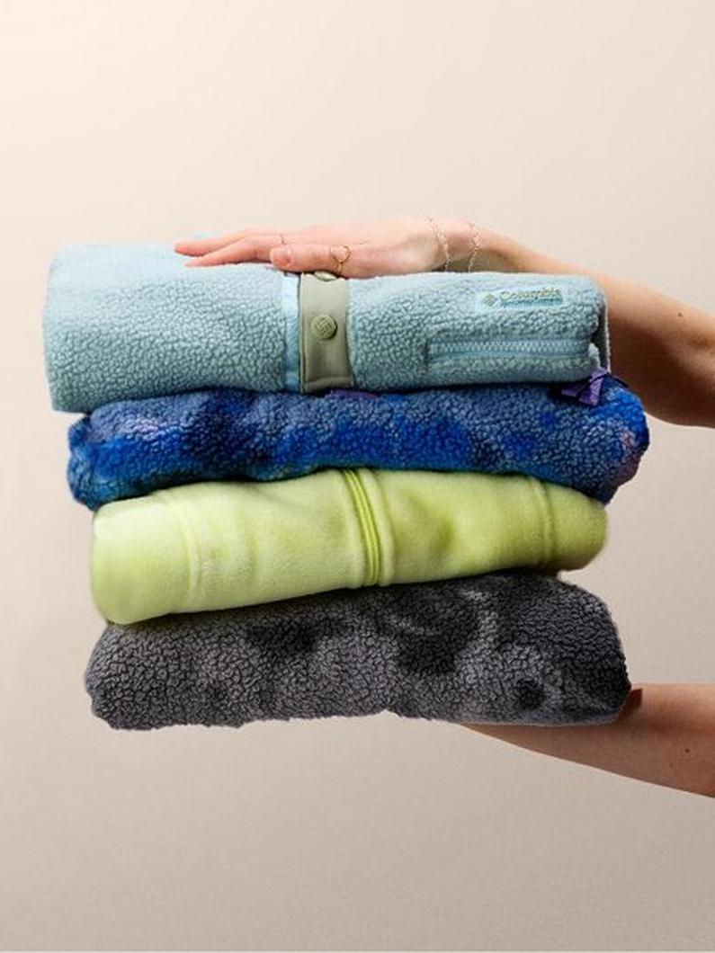 Hands holding a stack of four folded fleece jackets in light blue, royal blue, lime green, and gray.