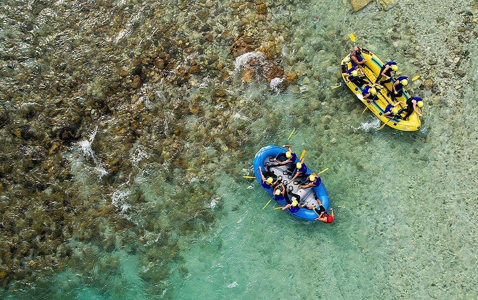 Two whitewater rafts, one yellow and the other blue, each carrying seven people, float side by side down a shallow stretch of river.