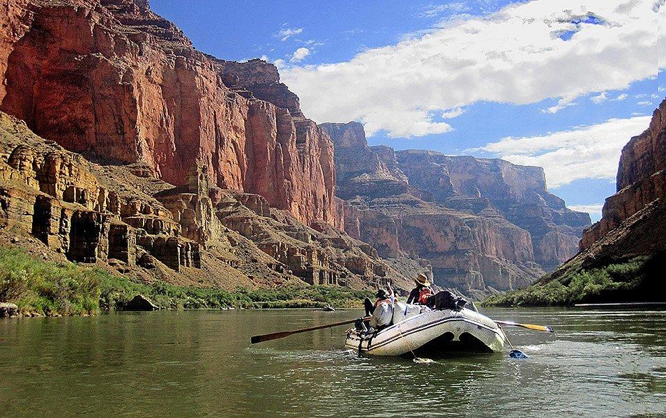 Paddlers wearing wide-brimmed hats navigate a calm stretch of flatwater in a whitewater raft, surrounded by towering canyon walls.