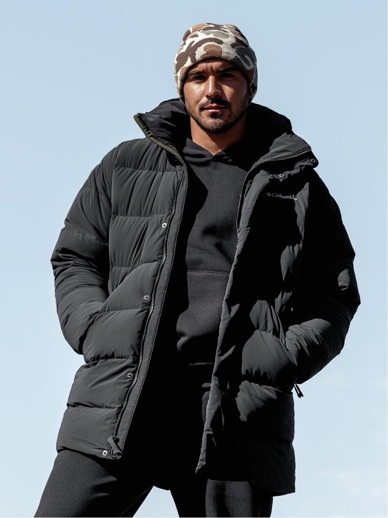Man wearing black puffer jacket over hoodie and camouflage beanie, standing outdoors.