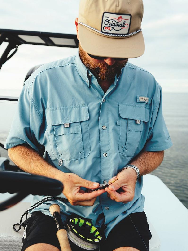 Bearded man in tan cap and light blue fishing shirt tying a lure on a boat with fishing reel nearby.