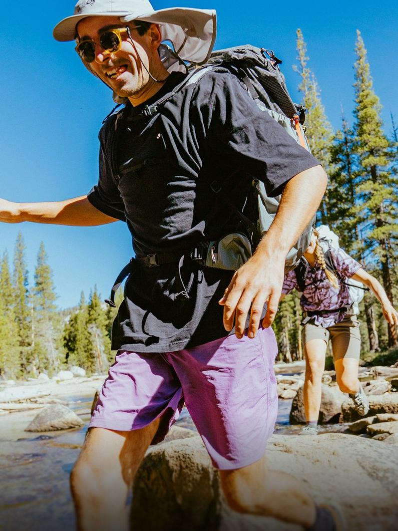 Man hiking in purple shorts, black tee, sunglasses, and white hat with backpack by a stream.