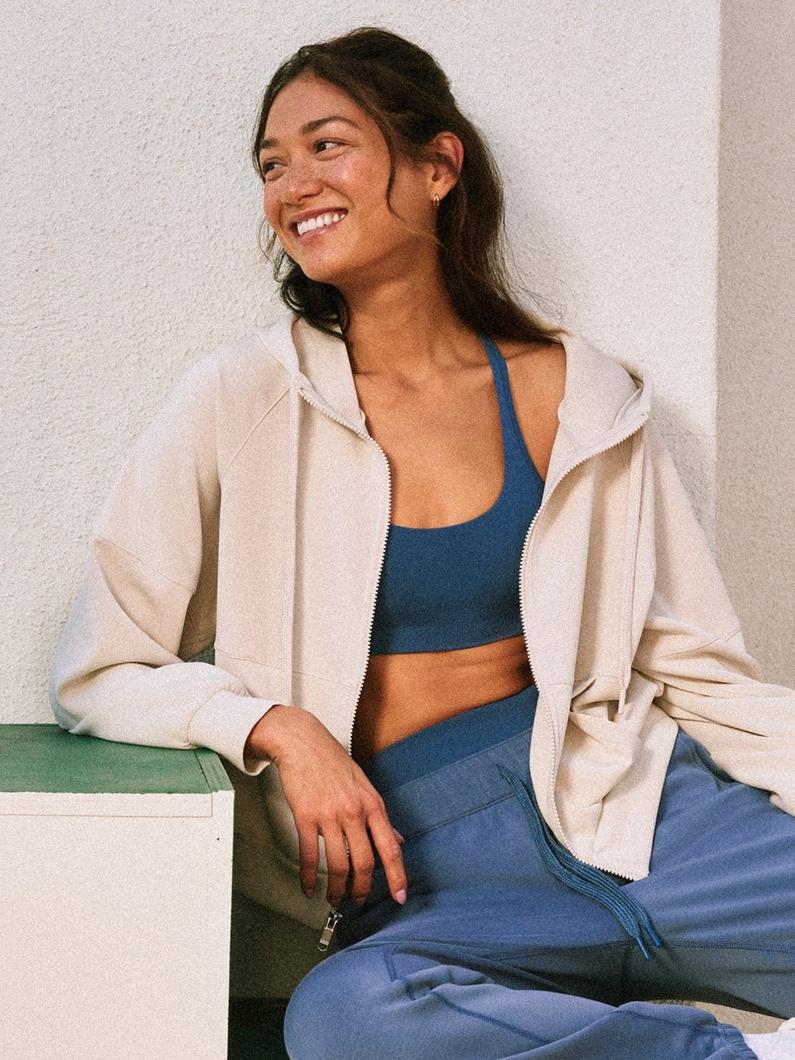 Woman sitting and smiling while wearing a blue sports bra, blue joggers, and beige zip-up hoodie.