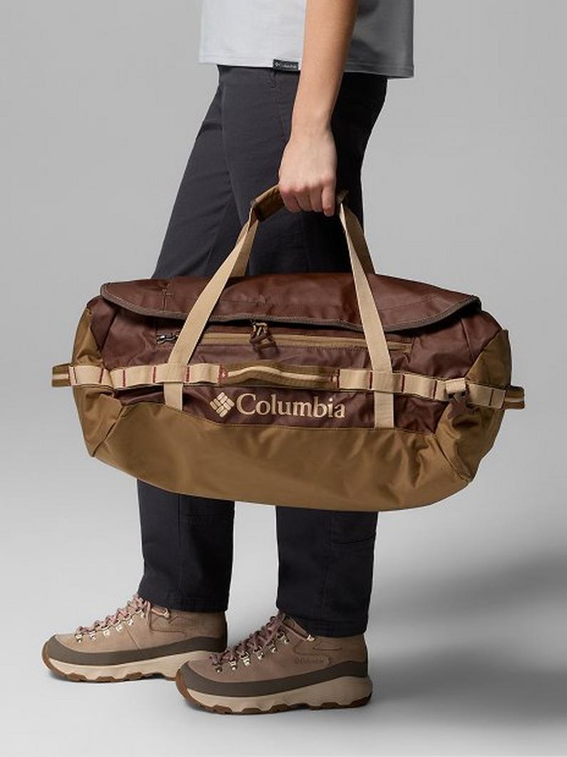 Person holding a brown and tan duffel bag with beige straps, wearing dark pants and hiking boots.