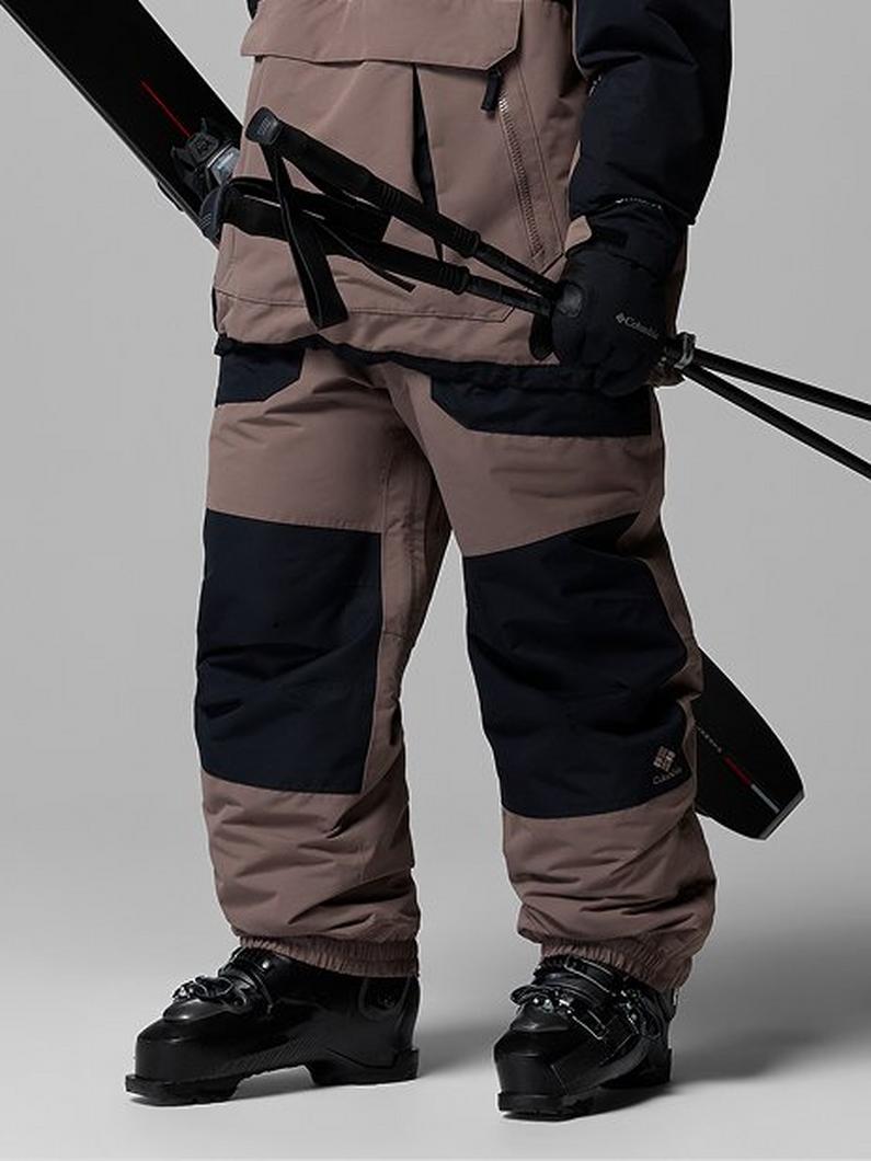 Person wearing tan and black ski pants with matching jacket, holding ski poles and skis in ski boots.