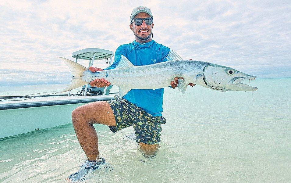 A man wearing PFG fishing gear kneeling in shallow water, while holding a large fish.  
