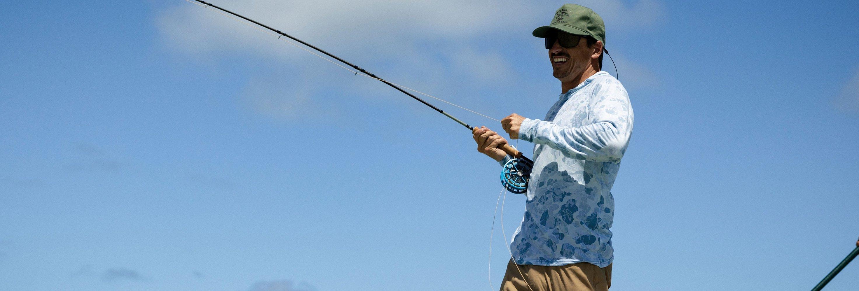 Man wearing green cap and fish-print performance shirt smiling while fly fishing with rod and reel.