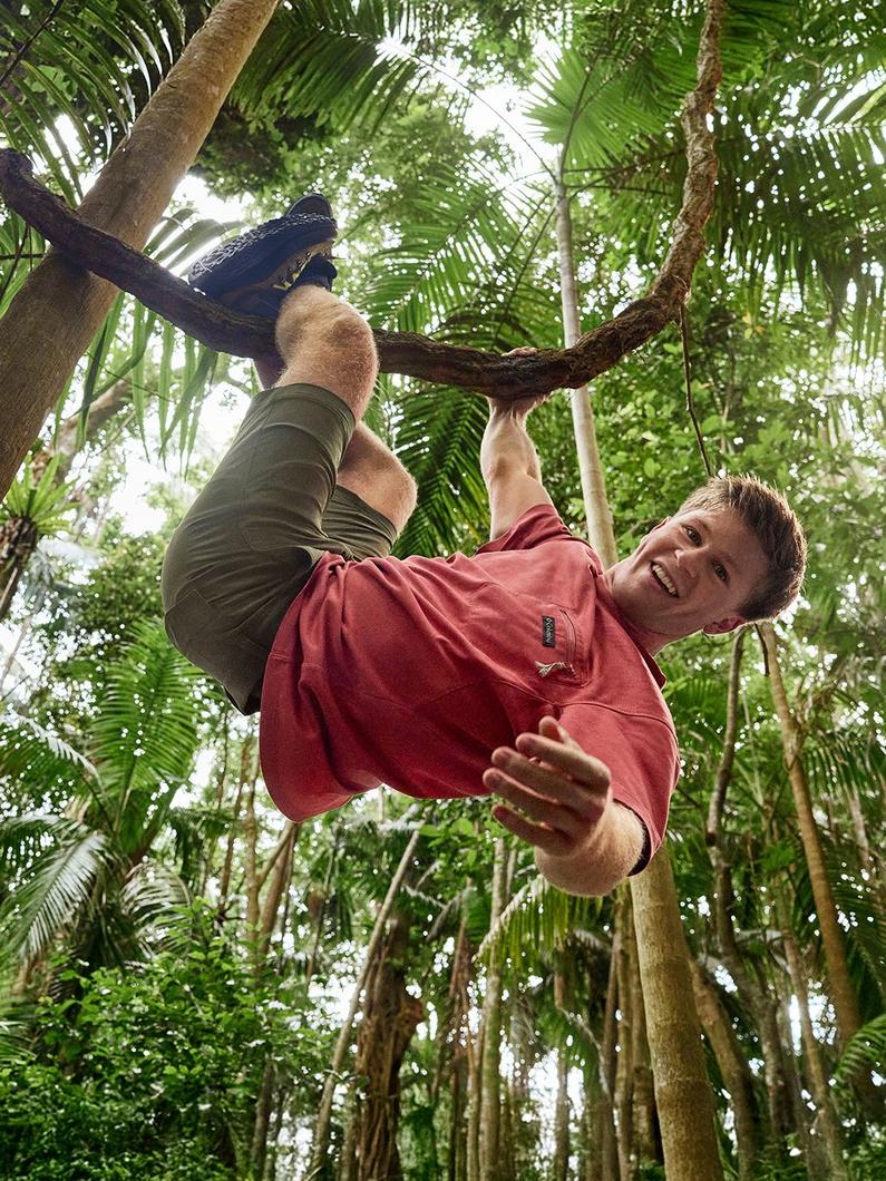 Man hanging from tree branch in tropical jungle, wearing red shirt and olive pants with hiking boots.