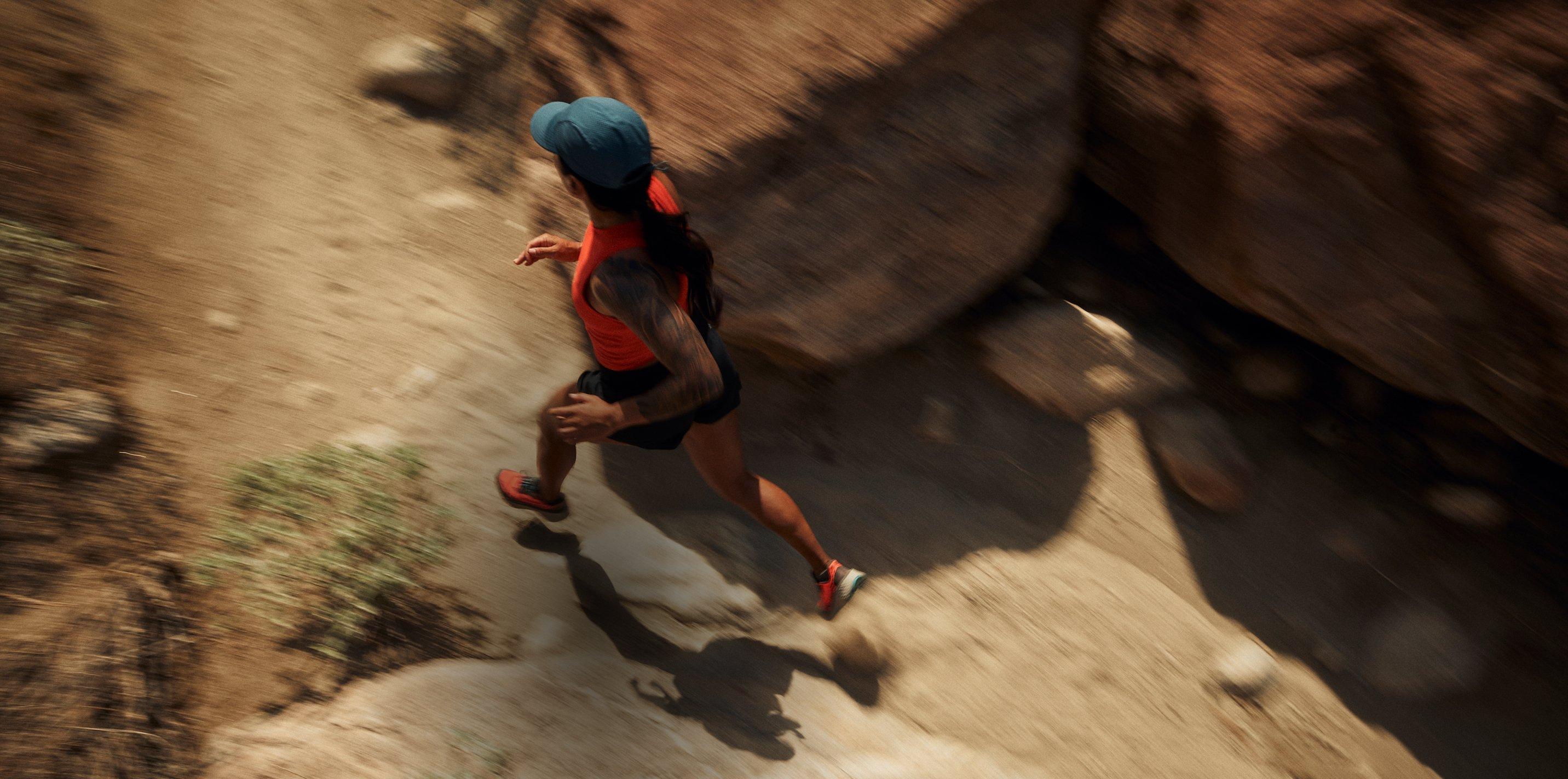 Person trail running in motion wearing blue cap, red shirt, black shorts, and orange running shoes.