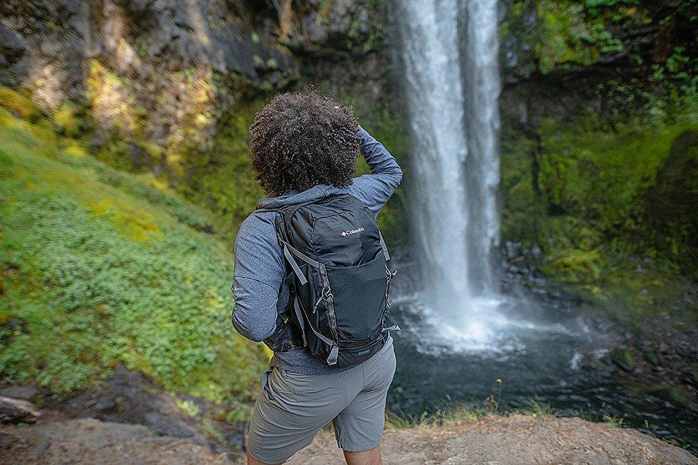 A backpacker pauses to look at a gorgeous cascading waterfall in the middle of a lush green forest. 