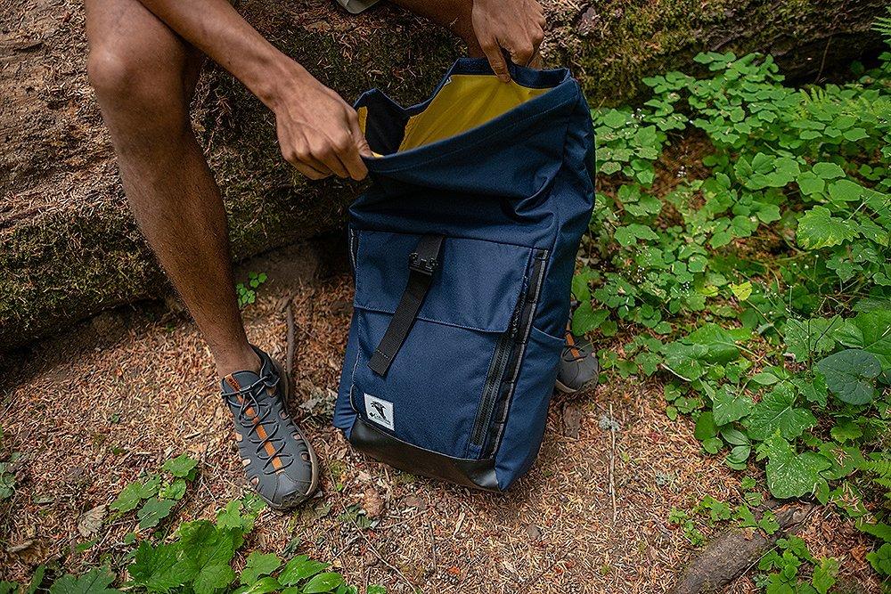 A close-up shot of a backpack on the ground in a scenic forest as a hiker’s hand digs through it. 