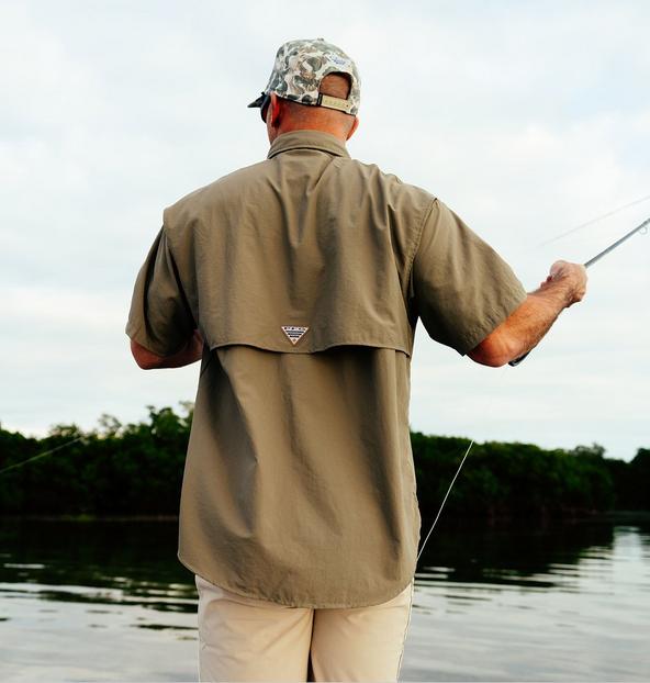 Man wearing tan vented fishing shirt and camouflage cap while holding fishing rod near water.